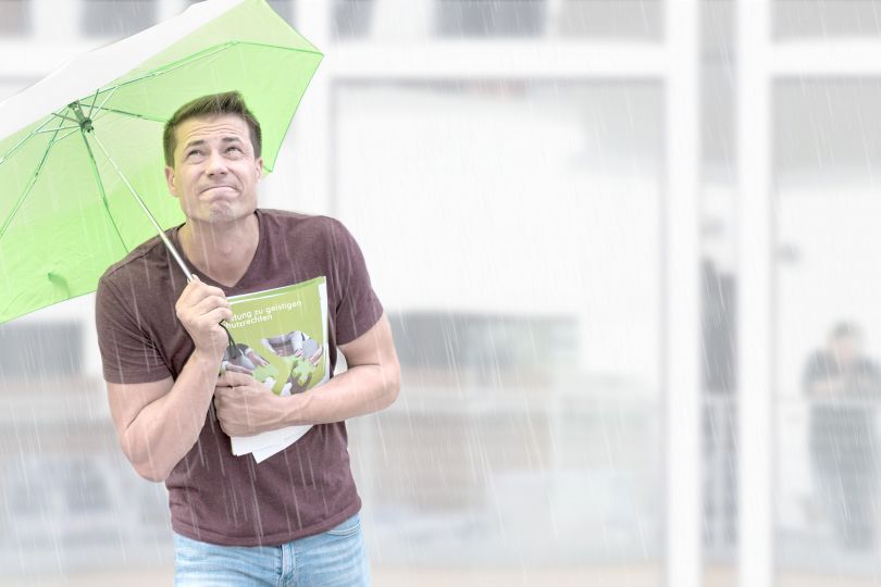 Ein junger Mann hält in einer Hand einen SFG-Folder zum Schutz von geistigem Eigentum, in der anderen einen grünen Schirm, um sich vor dem Regen zu schützen.