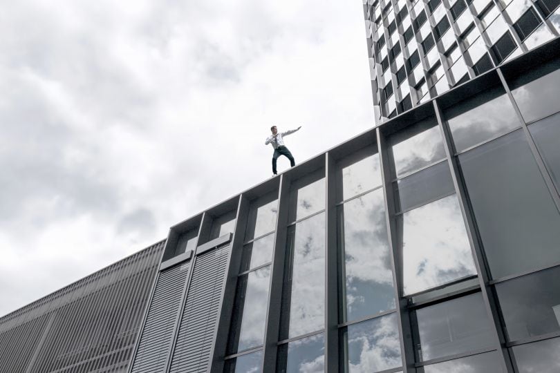 Ein junger Mann in Turnschuhen, Hemd und Krawatte steht auf einem Hochhaus in dynamischer Pose.