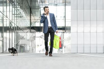 Ein Geschäftsmann spaziert mit einem Hund in der Leine, Einkaustaschen in der Hand telefonierend vor einer Glasfassade.