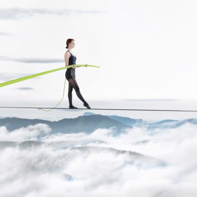 Frau balanciert auf Seil über Berglandschaft mit Wolken, in der Hand hält sie einen grünen Stab.