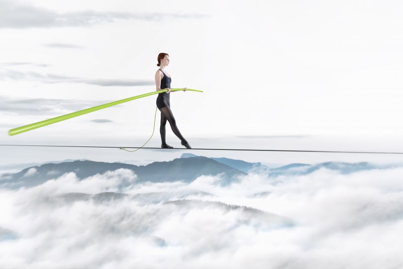 Frau balanciert auf Seil über Berglandschaft mit Wolken, in der Hand hält sie einen grünen Stab.