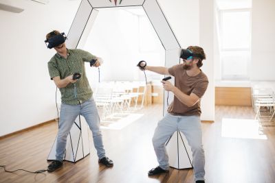 Zwei Männer mit Virtual-Reality-Brillen und Joysticks in der Hand vor weißem Bogen aus Papier.