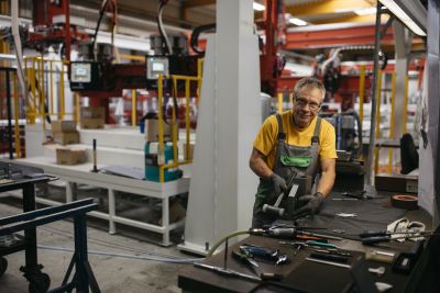 Mann mit Brille und Latzhose arbeitet mit Hammer in einer Produktionshalle