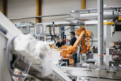 Zwei Männer stehen in Produktionshalle, im Vordergrund weiße und orange Maschinen