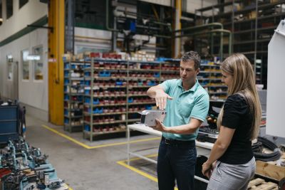 Florian Hampel hält in einer Produktionshalle Metallstück in der Hand und erklärt etwas Sigrid Merth