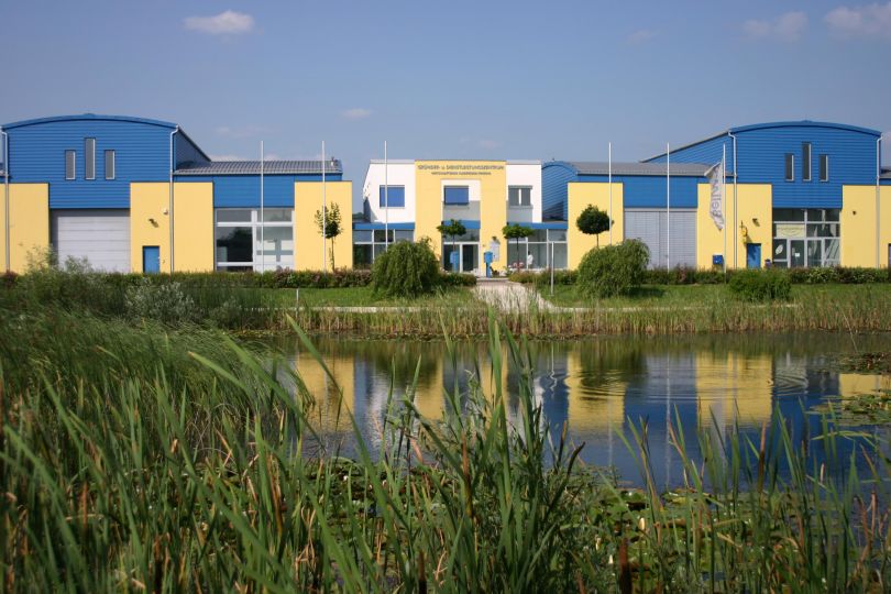 Gebäude in gelb, blau und weiß, davor ein Teich mit Schilf
