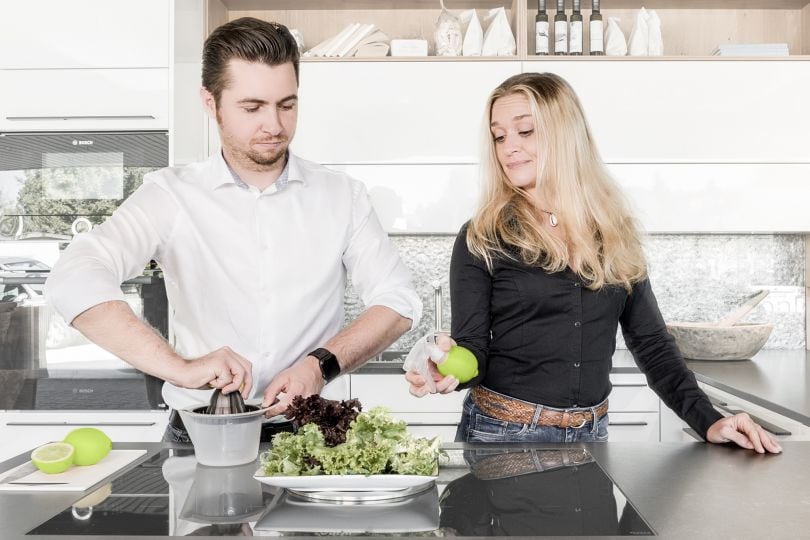 Ein Mann und eine Frau bereiten Salat zu. Der Mann presst eine Limette mit einer herkömmlichen Zitruspresse aus; die Frau hat eine Sprühvorrichtung in eine Limette gesteckt und zeigt dem Mann, wie es auf diese Weise einfacher geht.