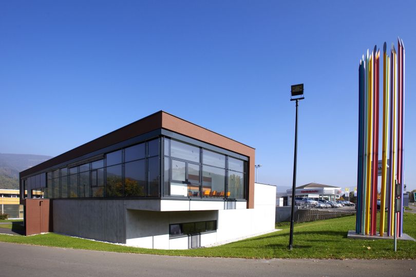 Gebäude in weiß, braun und Glas, gründe Wiese, blauer Himmel, rechts bunte Stäbe