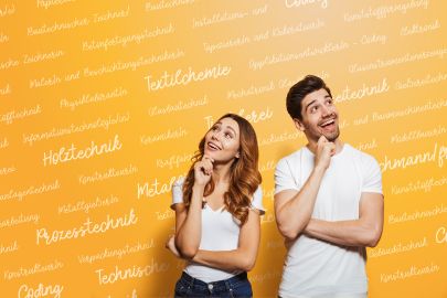 Eine junge Frau und ein junger Mann lächeln vor einer gelben Wand mit Schriftzügen technischer Berufe