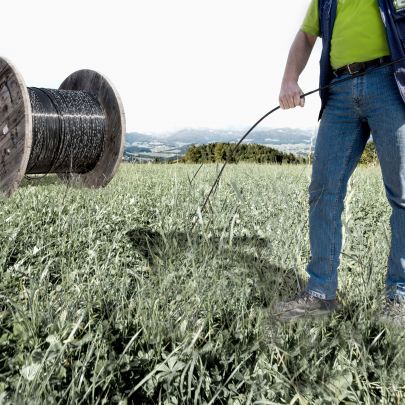 Mann auf Wiese hält Kabel in der Hand, im Hintergrund eine riesige Kabelrolle