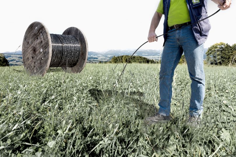 Mann auf Wiese hält Kabel in der Hand, im Hintergrund eine riesige Kabelrolle