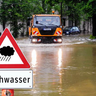 Hochwasser-Schild auf überfluteter Straße mit Einsatzfahrzeug