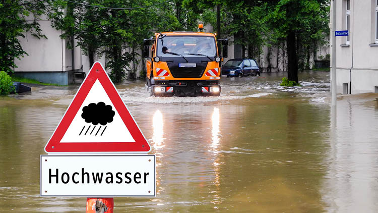 Hochwasser-Schild auf überfluteter Straße mit Einsatzfahrzeug